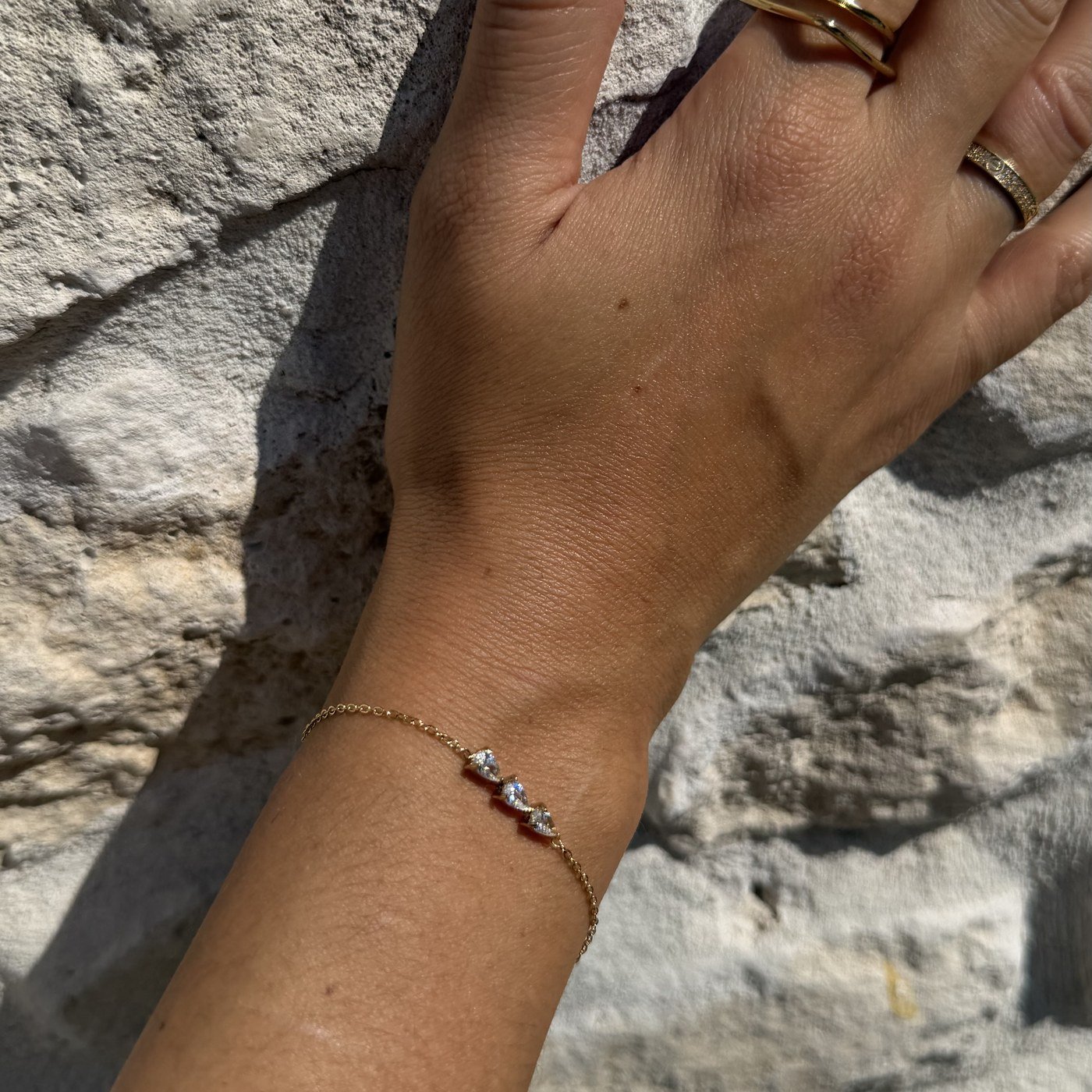 Bracelet Trilogie Doré 5 Bracelet trilogie doré au poignet, main posée sur un mur en pierre